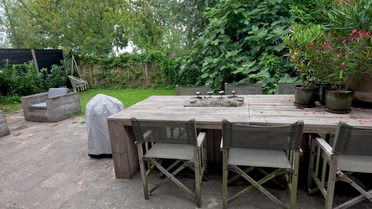 Camera slowly pans over outdoor patio with wooden furniture, lush greenery, and overcast daylight