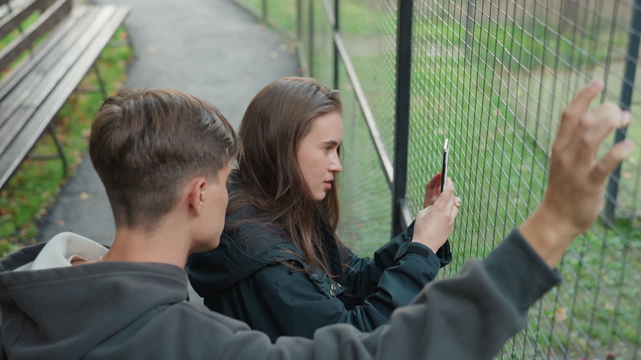 boy raising arm at metal fence while friend holds phone to grid, hoodieclad companion framing scene, tense protective gesture and overtheshoulder perspective, park path and bench visible, candid