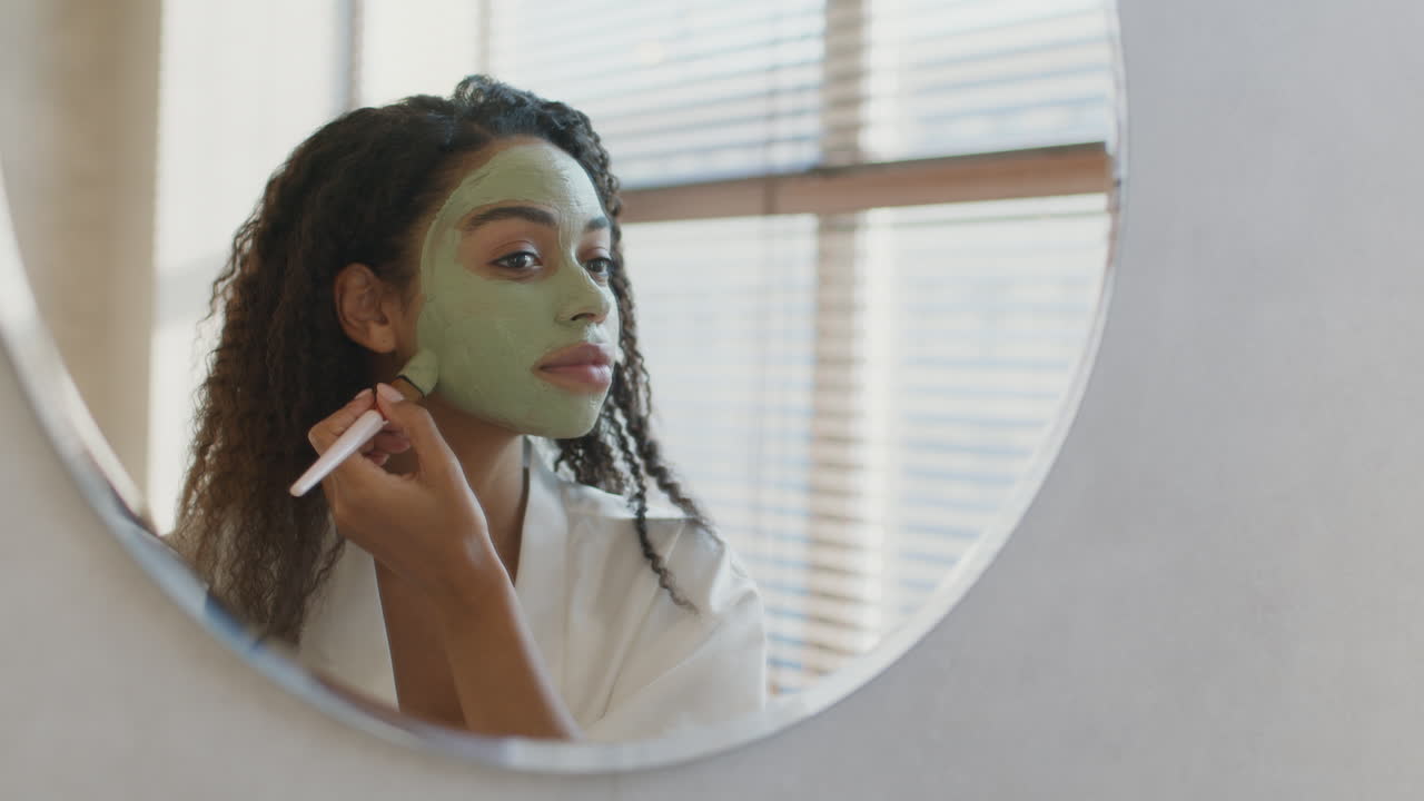 Woman applying a green face mask in front of a mirror