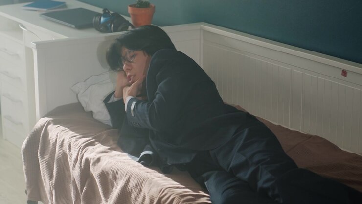 Serene individual sitting peacefully by window. Thoughtful man rests on bed with gentle sunlight illuminating. Contemplative Asian man relaxes on his bed near potted plant in soft morning light