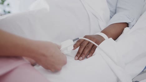 Hands-of-diverse-female-doctor-using-oximeter-on-finger-of-senior-female-patient-in-bed,-slow-motion