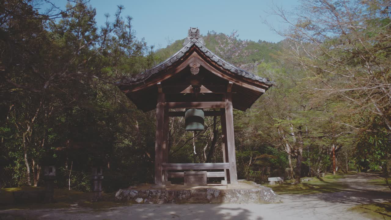 A traditional Shōrō bell building surrounded by lush woodland in Takaosan, Japan, showcasing tranquil natural beauty and cultural heritage on a clear day.