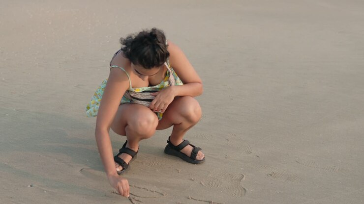 A young woman enjoying the beach, playfully writing on the sand with a small rock, creating sand art with joy