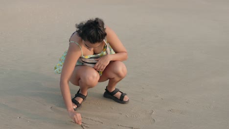 A-young-woman-enjoying-the-beach,-playfully-writing-on-the-sand-with-a-small-rock,-creating-sand-art-with-joy