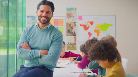 portrait of smiling male elementary school teacher working at desk in classroom with students