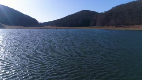 un sobrevuelo de drones cercano sobre un lago en las montañas, revela bosques y otros picos de montaña en la distancia