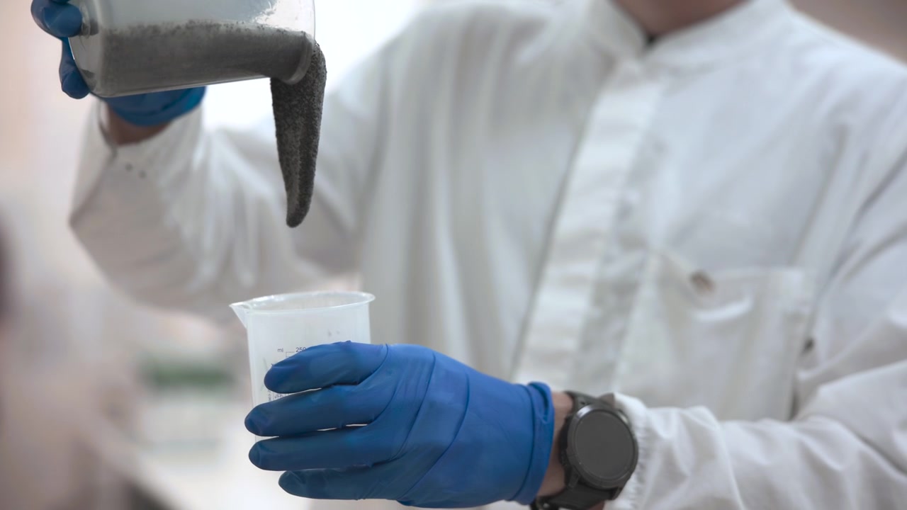 Close eye level view of gloved hands steadily pouring dark gray granular powder from tilted clear plastic container