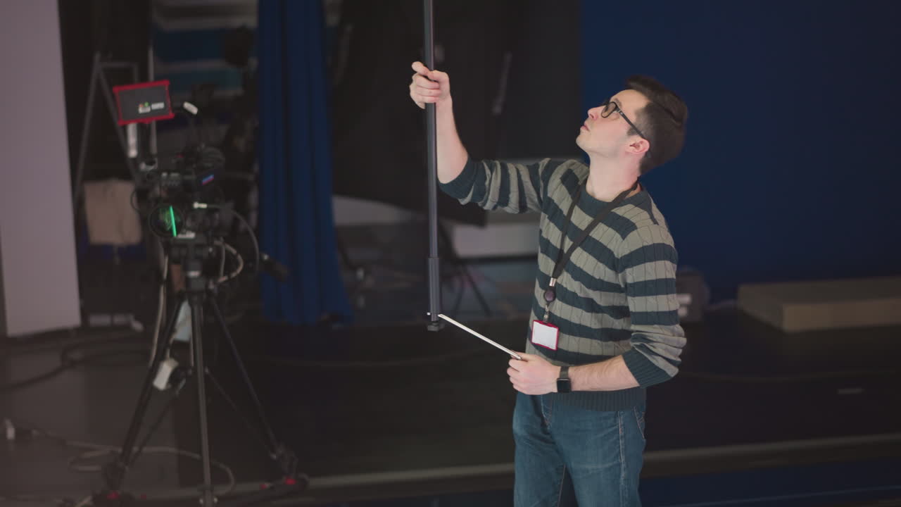 Man holding boom microphone, adjusting it in studio setting, broadcast or video production environment, focusing on camera equipment and crew, sound recording process in progress