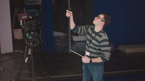 man holding boom microphone, adjusting it in studio setting, broadcast or video production environment, focusing on camera equipment and crew, sound recording process in progress