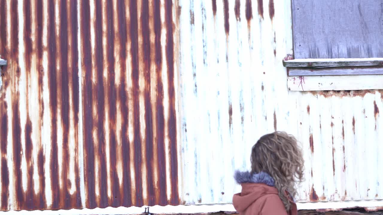 The slow pan right follows girl walking next to a rusty, abandoned house