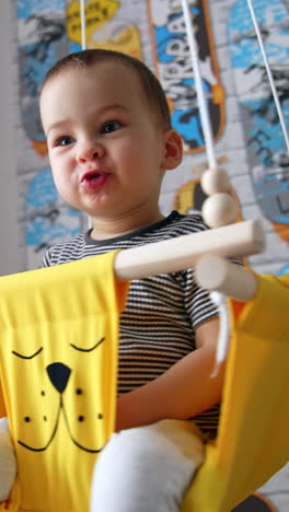 active cute caucasian baby boy sitting in a yellow swing. happy kid swinging at home. vertical video