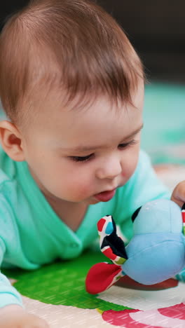 lovely baby boy in blue romper lies on colorful mat. cute kid playing with a toy, pulling it to his mouth and looking at it with interest. vertical video