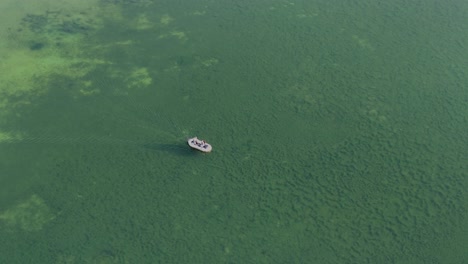 a inflatable boat slowly sails across a shallow lake with the bottom of plant life visible from an aerial drone