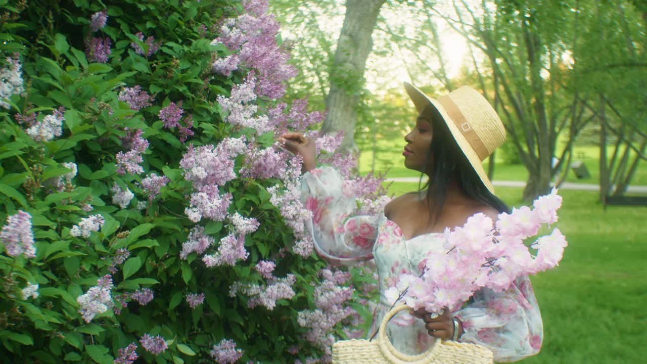 Gros plan d'une femme noire souriante, jouant avec des lilas et établissant un contact visuel.