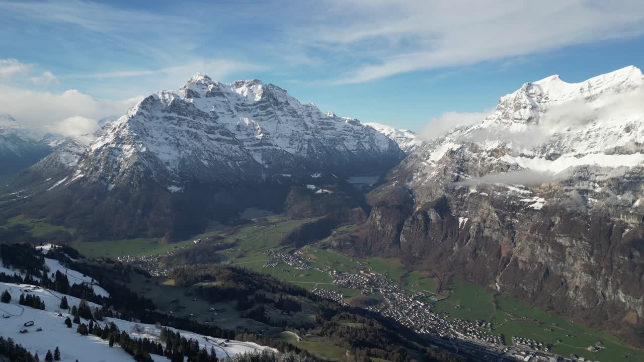 vista aérea de pájaro sobre el pueblo en la base del valle verde con nieve y nubes cubiertas de montañas