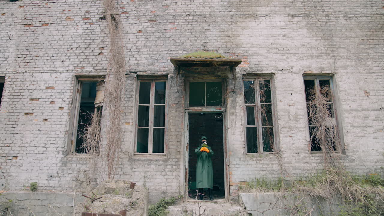 Human in infected zone. Ecologist in protective suit and gas mask coming out the abandoned building and holding flowers in hands.