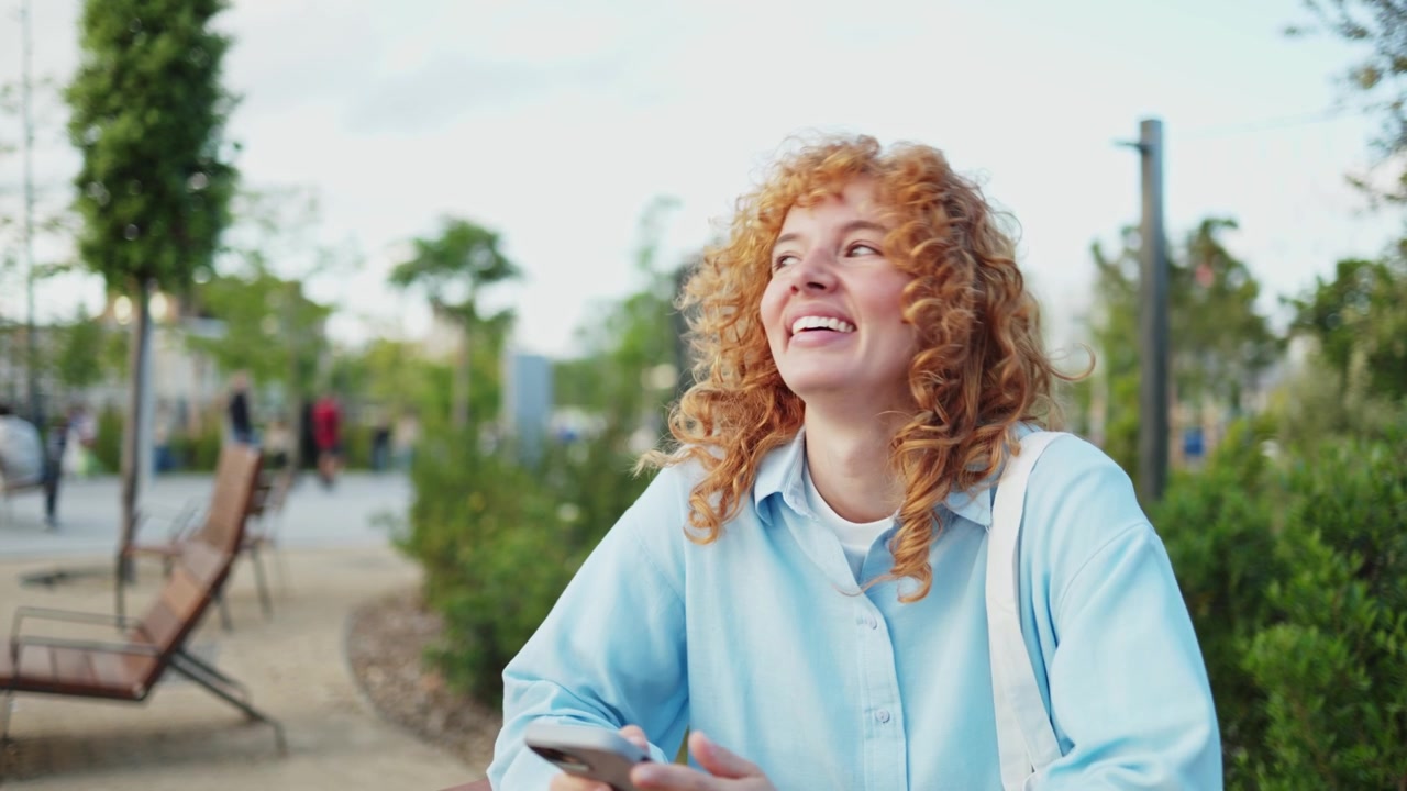 Blije jonge vrouw met krullend rood haar gebruikt smartphone in het park