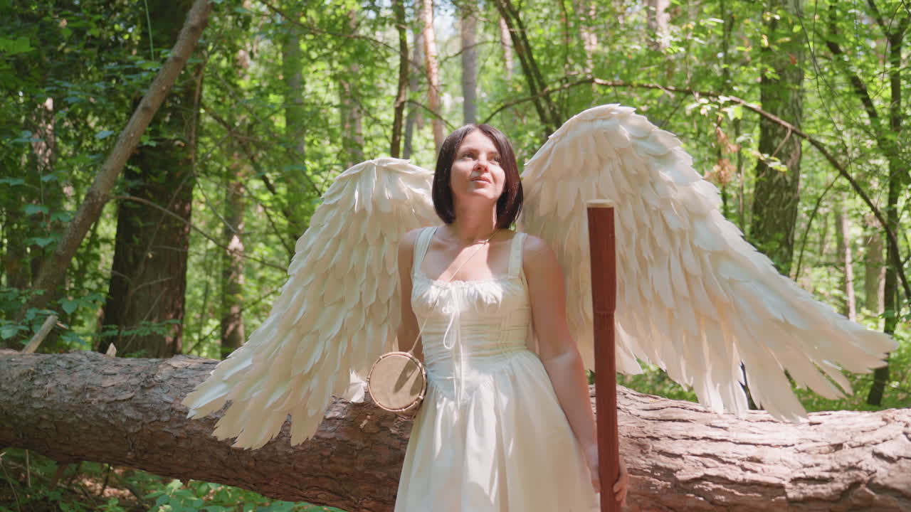 Signora vestita da angelo con grandi ali bianche e corto abito bianco appoggiata a un albero caduto, che tiene in mano un tamburo e un bastone di legno mentre sorride pacificamente sotto la luce del sole nella foresta, circondata dalla natura verde brillante