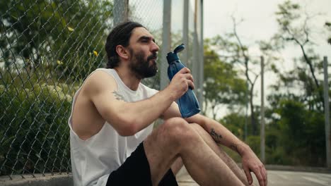 A-middle-aged-man-in-a-white-T-shirt-sits-on-a-basketball-court-near-the-fence,-rests-and-drinks-water-from-a-special-sports-bottle