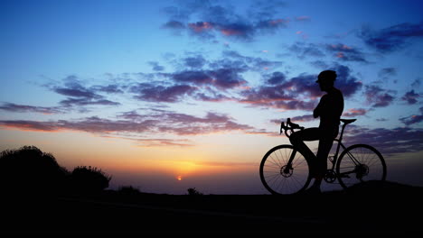 A-man-standing-atop-a-mountain-on-a-bicycle-takes-in-the-sunset's-beauty,-while-the-camera-smoothly-moves-on-a-Steadicam