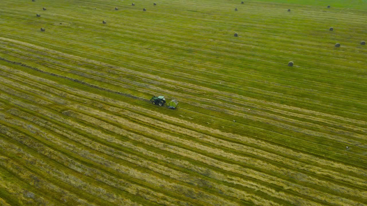 Aerial drone footage orbiting around a farm tractor vehicle that is making hay bale rolls with a machine attached in the back of the tractor on a green grassy farm field during cloudy day