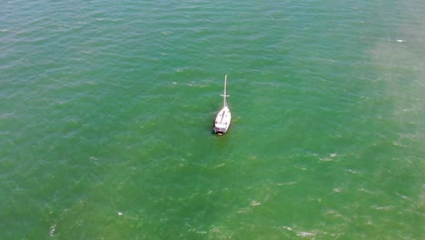 a small sailing boat being circled from above whilst it floats through small ripples of emerald green and demin blue water on a beautifully sunny and warm day