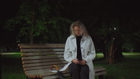 woman seated on wooden bench under streetlight at night using phone, surrounded by quiet green park with trees and empty pathway, black umbrella beside her