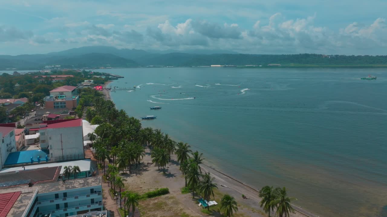 A dynamic drone shot captures the vast ocean buzzing with jet skis, a scenic shoreline, and buildings lining the coast.
