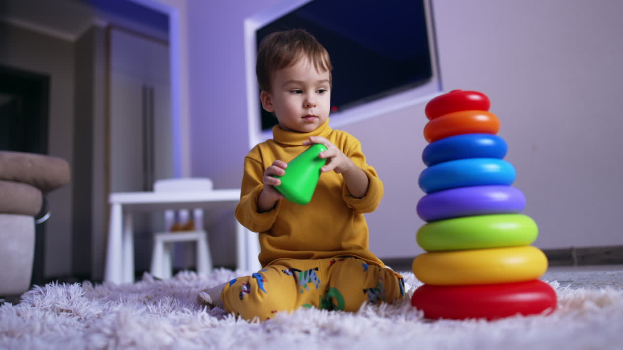 Serious baby boy sits on the floor playing with pyramid. Cute toddler in yellow sweater playing at home. Low angle view.