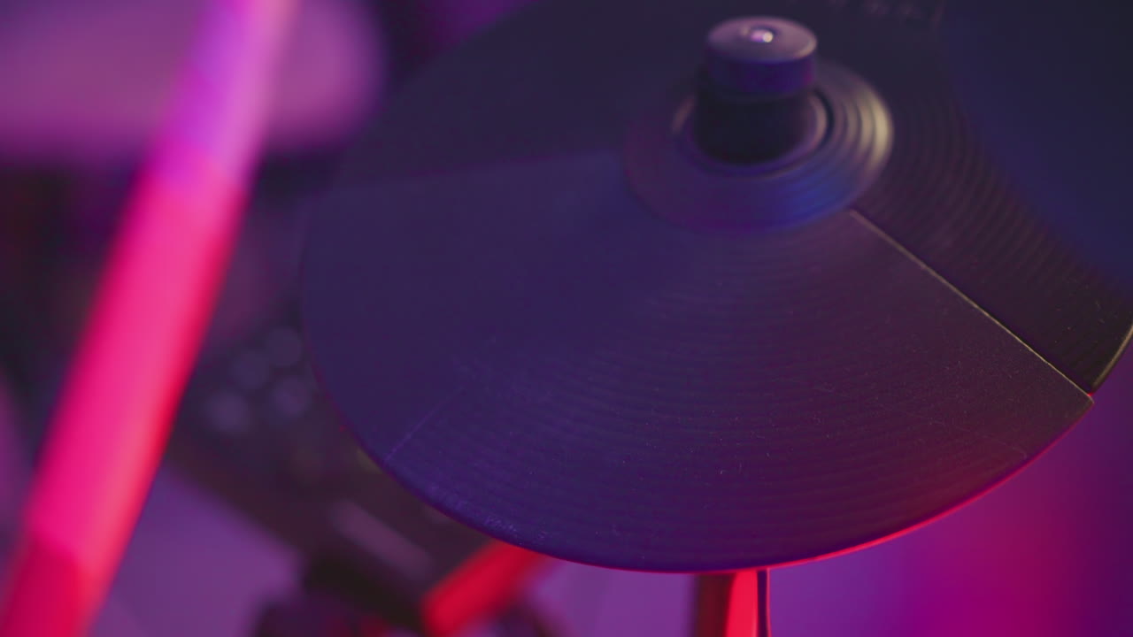 Close up of electronic cymbal being struck with drumstick under vibrant studio lighting, capturing percussion detail with motion and moody background during indoor drumming session