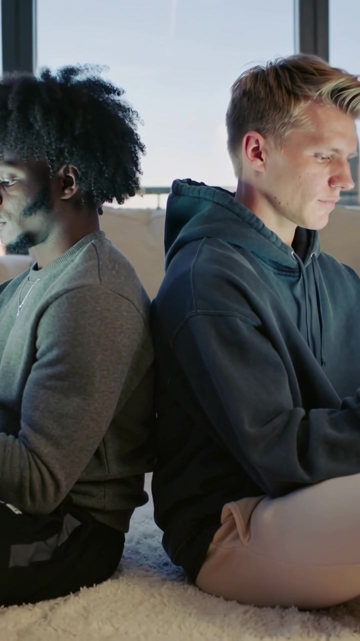 Vertical video: Sitting back-to-back, men scrolling then typing on phones at home, hoodie