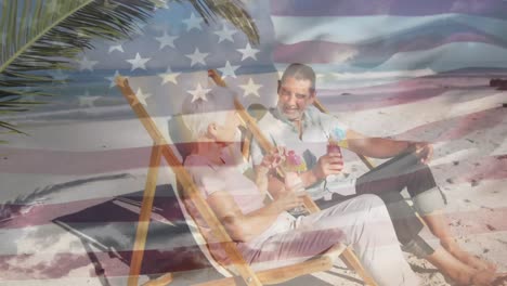 american flag waving against senior couple enjoying drinks sitting on deck chair at the beach