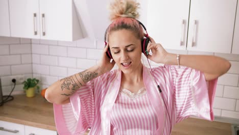 happy young woman with pink hair dancing in kitchen wearing pink pajamas and listening to music with headphones