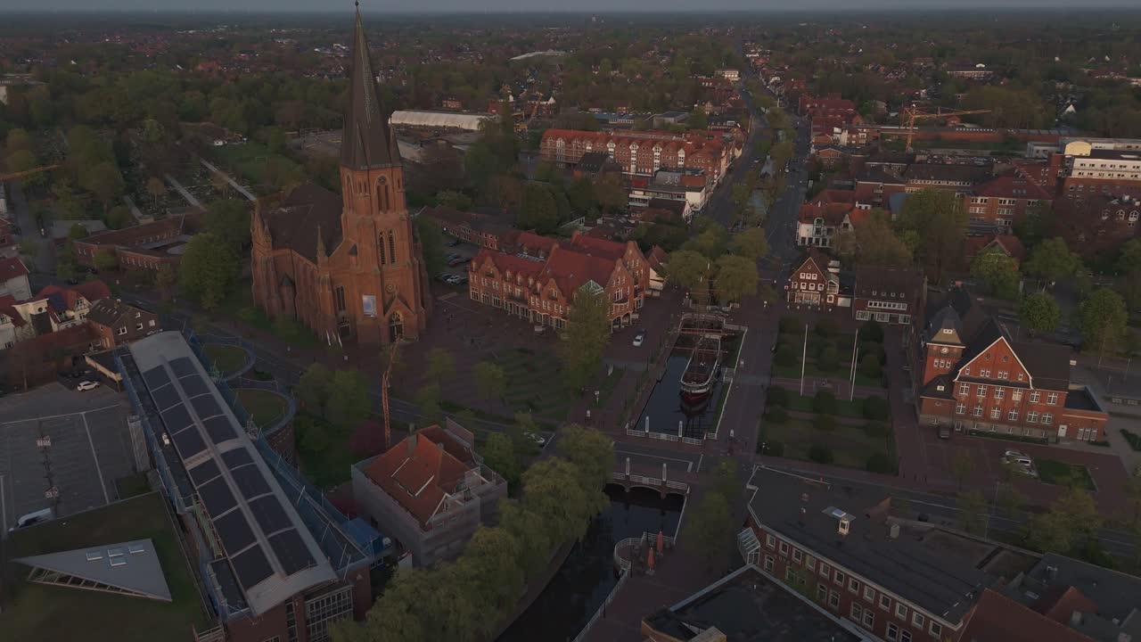 Aerial shot of Papenburg’s city center during golden hour, featuring St. Antonius Church and the historic ship Friederike von Papenburg on the main canal. Flying backward in soft evening light.