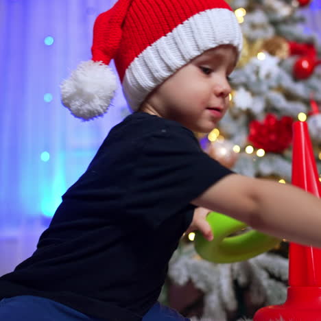cute caucasian toddler in red cap building a pyramid. christmas tree decorated for holiday at backdrop. low angle view.