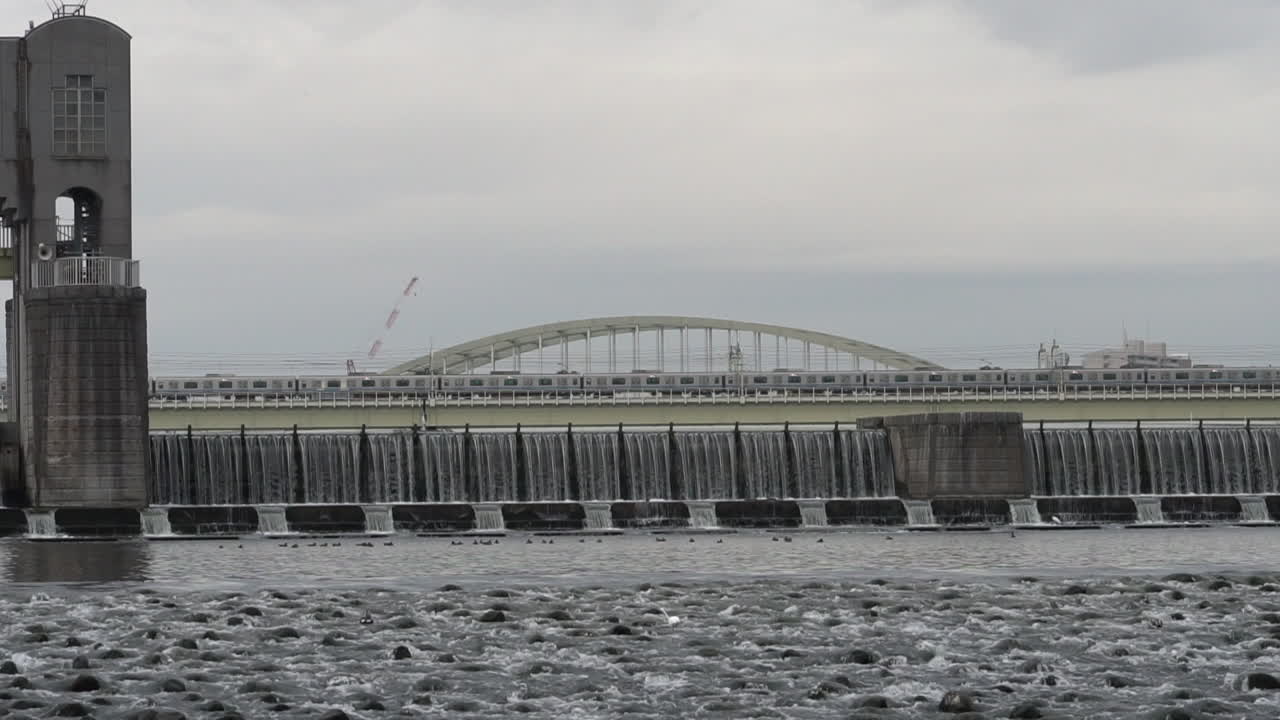 Water Flowing from Tama Ward Weir with Railway Running on the Background. Wild Birds Can be seen from the area. It was taken on a Gloomy day of Autumn Season at Tama Ward, Kawasaki, Japan