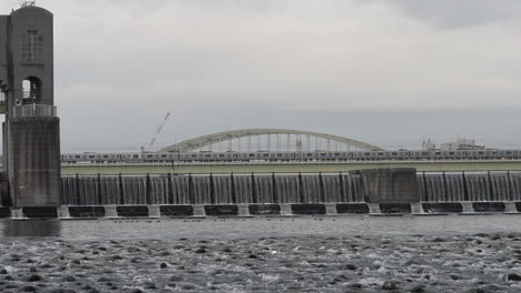 water flowing from tama ward weir with railway running on the background. wild birds can be seen from the area. it was taken on a gloomy day of autumn season at tama ward, kawasaki, japan