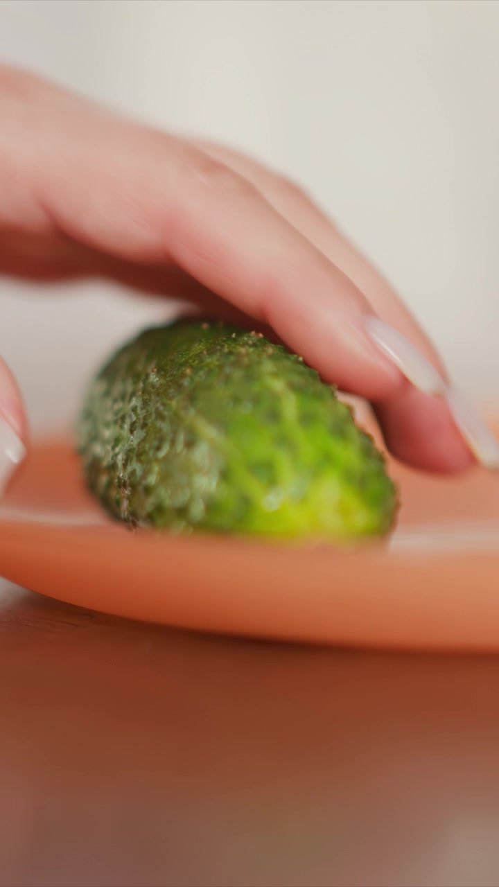 Una piccola verdura verde, probabilmente un pezzo di broccolo o un prodotto simile, poggia su un piatto arancione. Una mano appare brevemente per toccare il cibo prima che venga sollevato e rimosso dall'inquadratura. La scena si concentra da vicino.