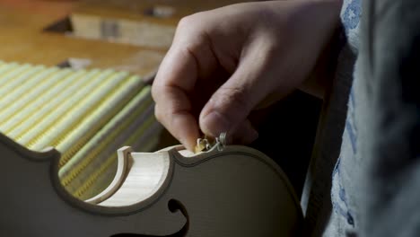 macro-close-up-of-artisan-luthier-maker-work-on-new-classic-handmade-raw-violin-smooth-edge-in-waist-and-purfling-cutter-knife-in-workbench-of-workshop-in-Cremona-Italy-4k-video