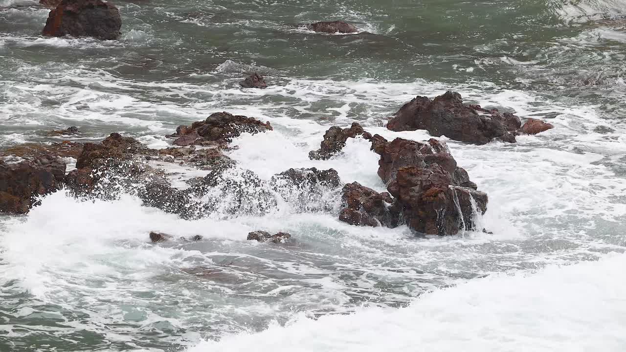 Dynamic waves crash against rugged rocks under natural light, capturing the raw power of the ocean