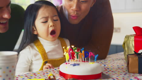 family with two dads celebrating daughter's birthday at home blowing out candles on cake