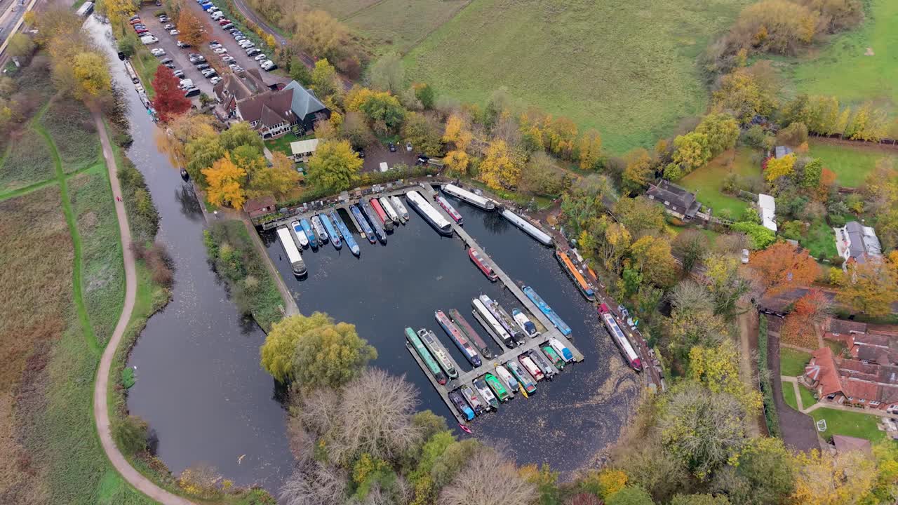 Drone orbits Harlow Marina in Essex. Autumn trees surround the calm, glass-like water, dotted with floating leaves and colourful canal boats, creating a serene and seasonal aerial view