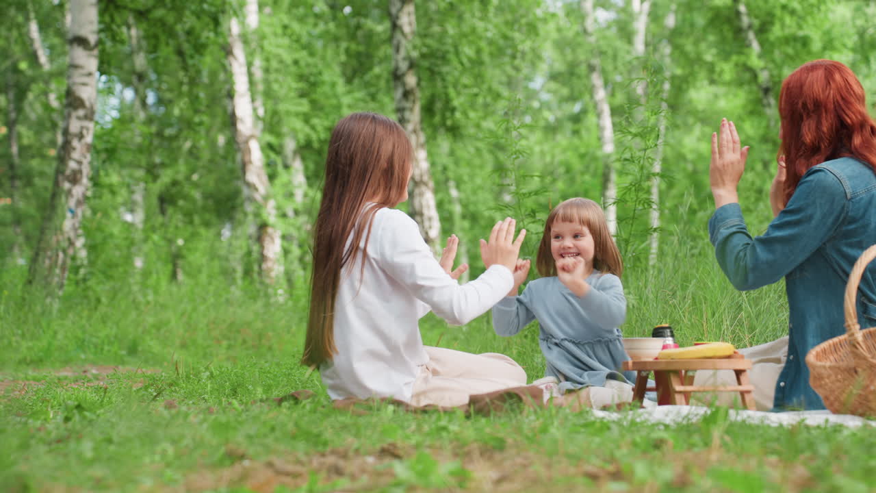 밝은 녹색 나무로 둘러싸인 야외 피크닉 장면에서 젊은 엄마가 매트 위에 앉아 두 딸과 함께 미소짓고 있어요. 막내딸이 엄마와 언니에게 하이파이브를 하려고 손을 뻗고 있어 즐거워 보여요.