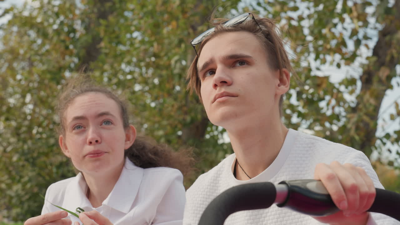 Teen caucasian siblings laughing in park, brother steady at stroller handle while sister giggles and sniffs leaf, warm sunlight through trees, upbeat playful family bonding, candid closeup capturing
