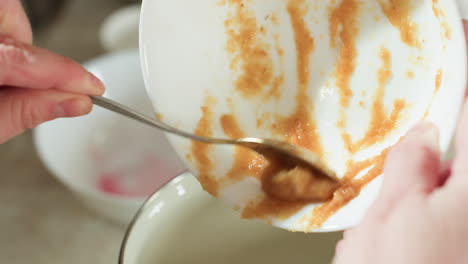 close up of person using spoon to transfer thick brown mashed ingredient from white bowl into stainless pot while cooking in kitchen, focusing on food texture