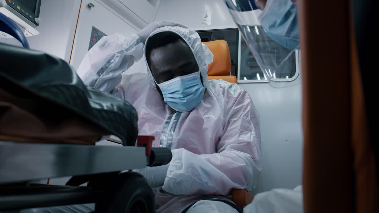 tired Black Paramedic Taking off Hazmat Suit Low Angle of African American Man in Hazmat Suit Taking off Mask and Face Shield while Riding Ambulance during Epidemic