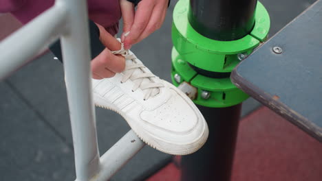 high angle shot of woman tying sneaker laces on iron exercise bar with foot perched on metal rail before outdoor workout session in urban fitness park near modern apartments with hoodie pulled up