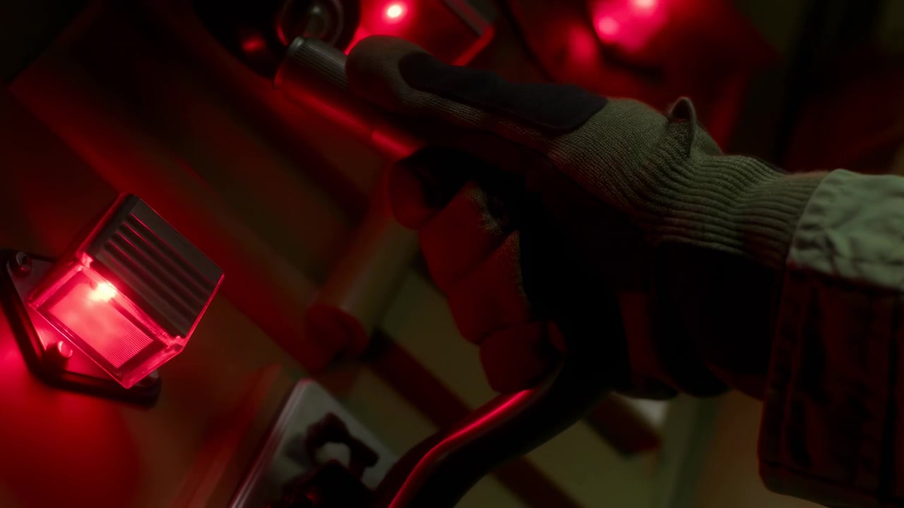 Una vista ravvicinata segue una mano guantata da lavoro che mantiene la presa su una maniglia di metallo testurizzata mentre le dita si muovono leggermente, con vivide lampade rosse che illuminano i vicini controlli e pannelli scuri e curvi.
