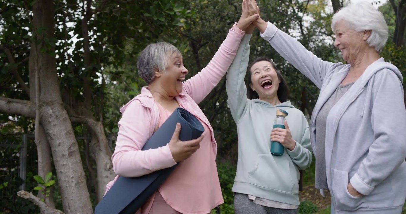 Mujeres diversas saludándose después de yoga en un parque, con esterilla azul, botella turquesa, levantando el brazo izquierdo para chocar los cinco.