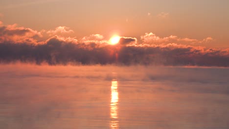 timelapse of the sun rising through clouds over a very cold lake with steam rising in the winter time as red yellow and red sun rays brighten the sky